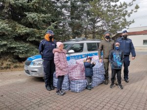 Na środku widoczny jest policyjny radiowóz przed którym leżą torby z prezentami i stoi dwóch policjantów w mundurach i jeden w ubraniu cywilnym (organizator przedsięwzięcia). Przed radiowozem stoi również trójka dzieci