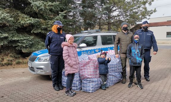 Na środku widoczny jest policyjny radiowóz przed którym leżą torby z prezentami i stoi dwóch policjantów w mundurach i jeden w ubraniu cywilnym (organizator przedsięwzięcia). Przed radiowozem stoi również trójka dzieci