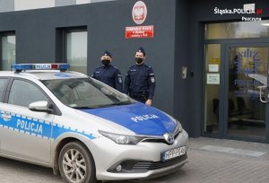 Na zdjęciu widoczni dwaj policjanci stojący obok radiowozu przed budynkiem Komisariatu Policji w Miedźnej. Pora dzienna