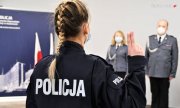 Nowo przyjęta policjantka podczas ślubowania trzyma w górze dwa palce, powtarzając słowa roty. Zdjęcie wykonane z tyłu, widoczny warkocz kobiety