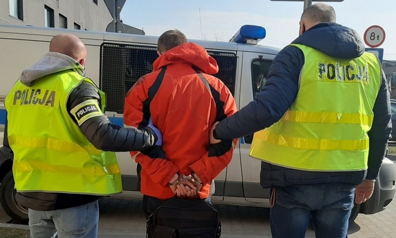 Dwóch policjantów, ubranych w zielone kamizelki policyjne trzyma zatrzymanego, wszyscy są odwróceni tyłem do aparatu. Zatrzymany ma założone kajdanki trzymane z tyłu, jest ubrany w czerwoną kurtkę. W tle widać radiowóz policyjny