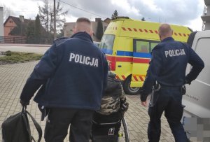 Dwaj umundurowani policjanci prowadza mężczyznę na wózku inwalidzkim - widok z tyłu. W tle widoczna jest karetka pogotowia.