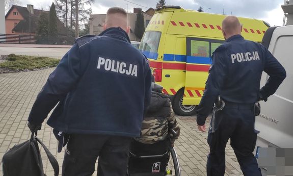 Dwaj umundurowani policjanci prowadza mężczyznę na wózku inwalidzkim - widok z tyłu. W tle widoczna jest karetka pogotowia