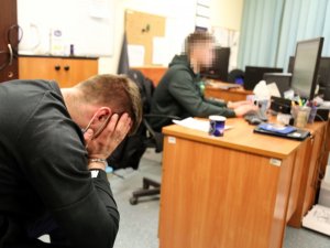 zatrzymany mężczyzna podczas przesłuchania siedzi na krześle w tle policjant przy biurku