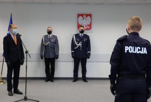 Umundurowani policjanci oraz osoby w garniturach stojący przy ścianie, nad nimi godło Polski