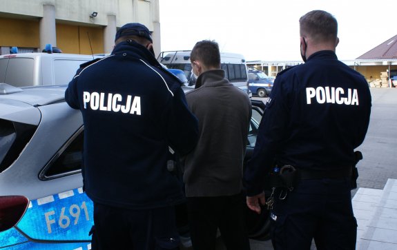 Umundurowani policjanci eskortują zatrzymanego do radiowozu.&quot;&gt;