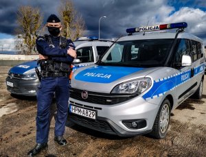 UMUNDUROWANY POLICJANT Z WYDZIAŁU PATROLOWO-INTERWENCYJNEGO BIAŁOSTOCKIEJ KOMENDY, KTÓRY STOI ZE SKRZYŻOWANYMI NA PIERSI RĘKAMI. POLICJANT STOI NA TLE DWÓCH RADIOWOZÓW POLICYJNYCH