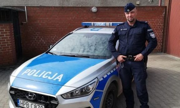 Na zdjęciu widać policjanta umundurowanego stojącego obok radiowozu na placu jednostki Policji. W tle widać garaże