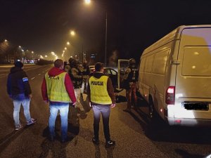 Przy lewych drzwiami samochodu typu bus koloru białego stoją 4  policjanci pododdziału kontrterrorystycznego ubrani w moro oraz taktyczne czarną kamizelkę, z boku samochodu stoją dwaj policjanci, które mają założone  kamizelki odblaskowe z napisem policja&quot;&gt;