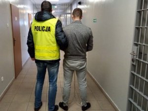 w areszcie policjant ubrany w kamizelkę odblaskowa z napisem POLICJA prowadzi zatrzymanego który ma założone kajdanki na ręce trzymane z przodu oraz na stopy, z prawej strony widać kratę na oknie, z lewej drzwi do pomieszczeń