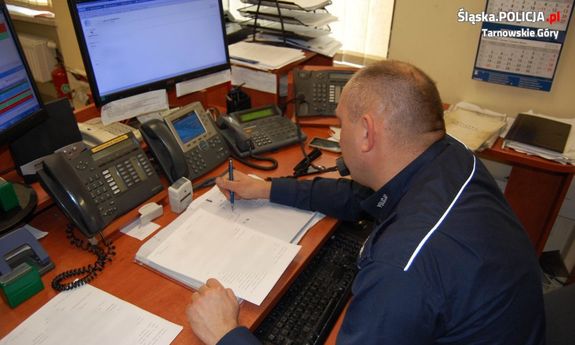 Zdjęcie kolorowe. Na fotografii widać siedzącego przy biurku policjanta. Mundurowy rozmawia przez telefon. Na biurku widać monitory komputerowe oraz aparaty telefoniczne