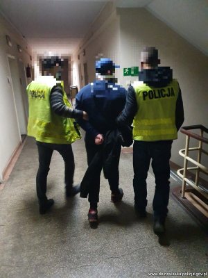 Na zdjęciu widać dwóch policjantów w kamizelkach odblaskowych, którzy prowadzą na schody zatrzymanego mężczyznę