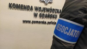 na zdjęciu widoczny jest napis na ścianie: Komenda Wojewódzka w Gdański i fragment adresu www.pomorska.policja. oraz rękaw policjanta, który ma założoną niebieską opaskę z białym napisem: negocjator