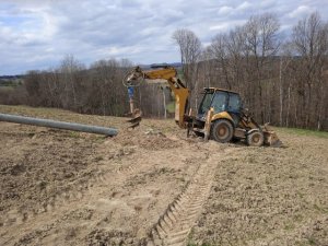 Koparka CAT z zamontowanym na ramieniu świdrem. Obok leży betonowy słup energetyczny