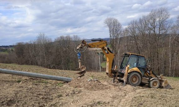 Koparka CAT z zamontowanym na ramieniu świdrem. Obok leży betonowy słup energetyczny