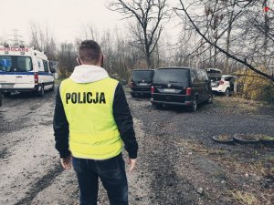 Policjant ubrany w kamizelkę z napisem policja stoi na pierwszym planie, a za nim znajdują się samochody biorące udział w pościgu