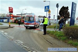 Policjant wykonujący czynności na miejscu zdarzenia drogowego, w tle karetka pogotowia i radiowóz