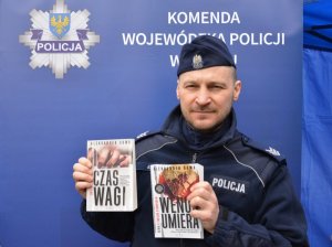 Umundurowany policjant na tle ścianki z napisem Komenda Wojewódzka Policji w Opolu trzyma dwie książki w ręku