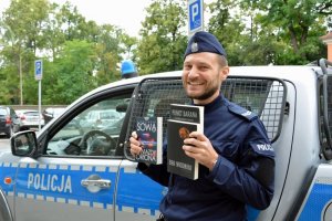 Umundurowany policjant przy radiowozie w ręku trzyma trzy książki