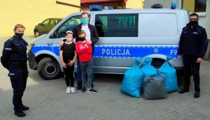 policjant i policjantka w mundurze, radiowóz oznakowany bus oraz mężczyzna z dwoma małymi dziewczynkami i foliowe worki w których znajdują się nakrętki