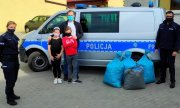 policjant i policjantka w mundurze, radiowóz oznakowany bus oraz mężczyzna z dwoma małymi dziewczynkami i foliowe worki w których znajdują się nakrętki