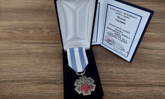 Na biurku leży w pudełku medal w kształcie gwiazdy, a środku czerwony krzyż i kwiat lilii oraz biało- niebieska szarfa. Z tyłu postawiona książeczka z tekstem ZASŁUŻONY HONOROWY DAWCA KRWI Marcin Choiński odznaczony/a został/a odznaką HONOROWY DAWCA KRWI ZASŁUŻONY DLA ZDROWIA NARODU. Na dole pieczątka: Z upoważnienia MINISTRA ZDROWIA PODSEKRETARZ STANU Sławomir Gadowski oraz własnoręczny podpis. W prawym dolnym rogu: Minister Zdrowia