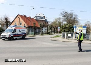 Policjant kieruje ruchem na skrzyżowaniu. W tle przejeżdżający ambulans medyczny.&quot;&gt;