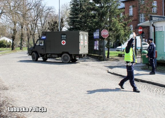 Policjant kieruje ruchem na skrzyżowaniu. W tle przejeżdżający ambulans medyczny.&quot;&gt;