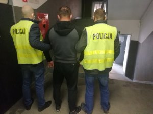 Policjanci ubrani w żółte kamizelki z napisem Policja prowadzą zatrzymanego. W tle klatka schodowa w budynku Komendy Miejskiej Policji w Łodzi