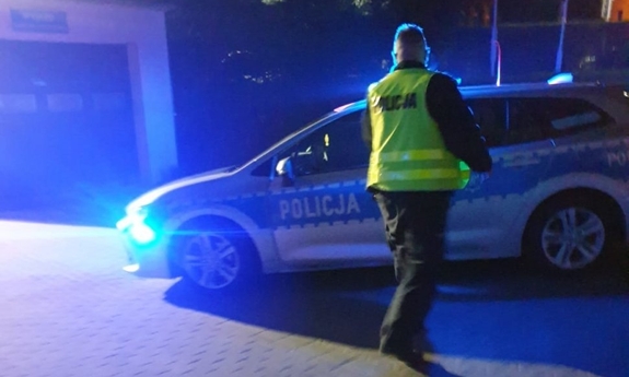 Zdjęcie przedstawia policjanta zmierzającego do radiowozu z włączonymi sygnałami świetlnymi