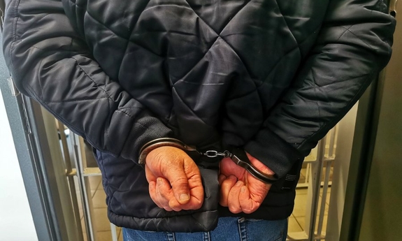 Ręce zatrzymanego mężczyzny, trzymane z tyłu z zapiętymi kajdankami. Stoi przy kracie pomieszczenia dla osób zatrzymanych