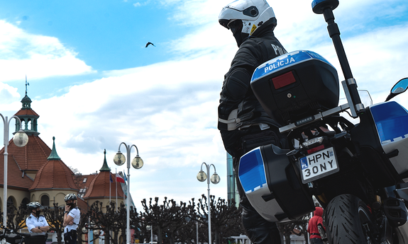 policjant przy motocyklu, w tle budynek i dwie policjantki