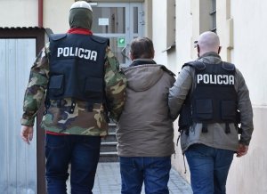 na zdjęciu dwóch mężczyzn w czarnych kamizelkach z napisem policja prowadzi mężczyznę w brązowej kurtce. W tle przeszklone drzwi do budynku
