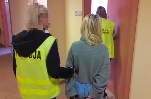 policjantki ubrane w kamizelki odblaskowe z napisem na plecach policja, wprowadzają do pokoju przesłuchań młodą kobietę która na rękach trzymanych z tyłu ma nałożone kajdanki