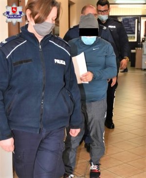 zatrzymany mężczyzna na korytarzu budynku, ma na rękach i nogach kajdanki obok idą policjanci