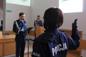 umundurowany  policjant przed mikrofonem, nowo przyjęta policjantka z ręka uniesioną do góry składa ślubowanie, z tyłu widoczny policjant za mównicą