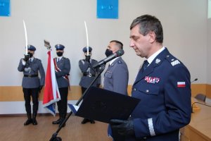 umundurowany policjant przed mikrofonem, z tyłu widoczny inny umundurowany funkcjonariusz i poczet sztandarowy