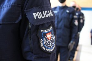 na pierwszym planie widoczny rękaw policyjnego munduru z naszywką, z tyłu dwaj nowo przyjęci policjanci