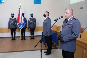 umundurowany policjant przed mikrofonem, z tyłu widoczny inny umundurowany funkcjonariusz i poczet sztandarowy