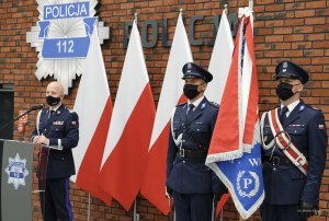 umundurowany policjant za mównica, obok poczet sztandarowy, z tyłu ustawione biało - czerwone flagi a an ścianie gwiazda policyjna i napis Policja