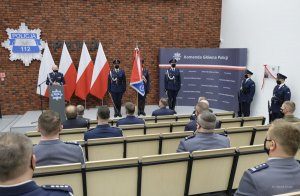 umundurowany policjant za mównica, obok poczet sztandarowy, z tyłu ustawione biało - czerwone flagi a an ścianie gwiazda policyjna i napis Policja, przed nimi, tyłem na zdjęciu zgromadzeni słuchacze
