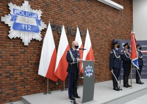 umundurowany policjant za mównica, obok poczet sztandarowy, z tyłu ustawione biało - czerwone flagi a an ścianie gwiazda policyjna i napis Policja