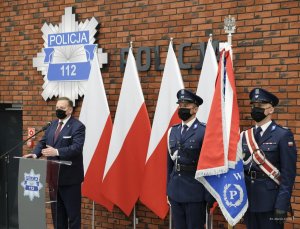 mężczyzna w garniturze za mównicą, obok poczet sztandarowy, z tyłu ustawione biało - czerwone flagi, na ścianie gwiazda policyjna i napis Policja