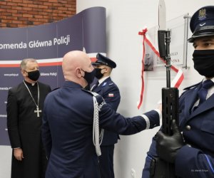 umundurowany policjant zrywa biało-czerwona wstęgę z tablicy, za nim stoi biskup w sutannie, po obu stronach tablicy policjanci z bagnetami