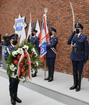 policjant trzyma przed sobą wieniec z kwiatów, obok poczet sztandarowy, z tyłu ustawione biało - czerwone flagi a an ścianie gwiazda policyjna i napis Policja