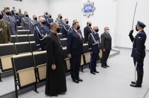 mężczyźni  w mundurach i po cywilnemu stoją na przeciwko umundurowanego policjanta, który trzyma w dłoni  uniesioną do góry szablę