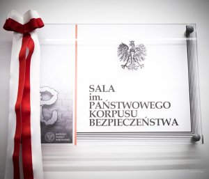 tablica z napisem: Sala konferencyjna im. Państwowego Korpusu Bezpieczeństwa, z boku, z lewej strony zwisa biało - czerwona wstęga