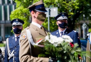 na pierwszym planie żołnierz niosący wieniec kwiatów, w tle dwaj policjanci