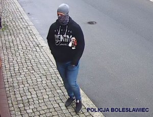 Na zdjęciu widoczna jest osoba średniego wzrostu, krępej budowy ciała, ubrana w ciemną odzież i obuwie sportowe. Na czarnej bluzie z kapturem znajdują się jasne napisy. Na twarzy ma założony różnobarwny komin a na głowie szarą czapkę. Osoba trzyma ręce w kieszeniach