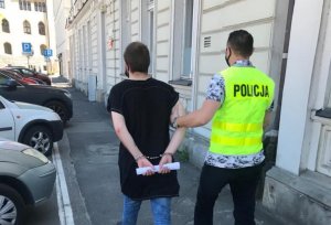 Policjant prowadzi zatrzymanego po chodniku. Z lewej strony widoczne zaparkowane pojazdy, a z prawej budynek